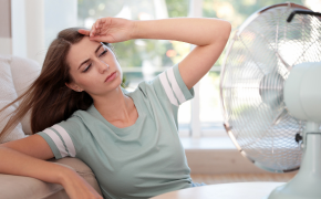 Frau sitzt mit wehenden langen Haaren auf Sofa vor einem Ventilator. 